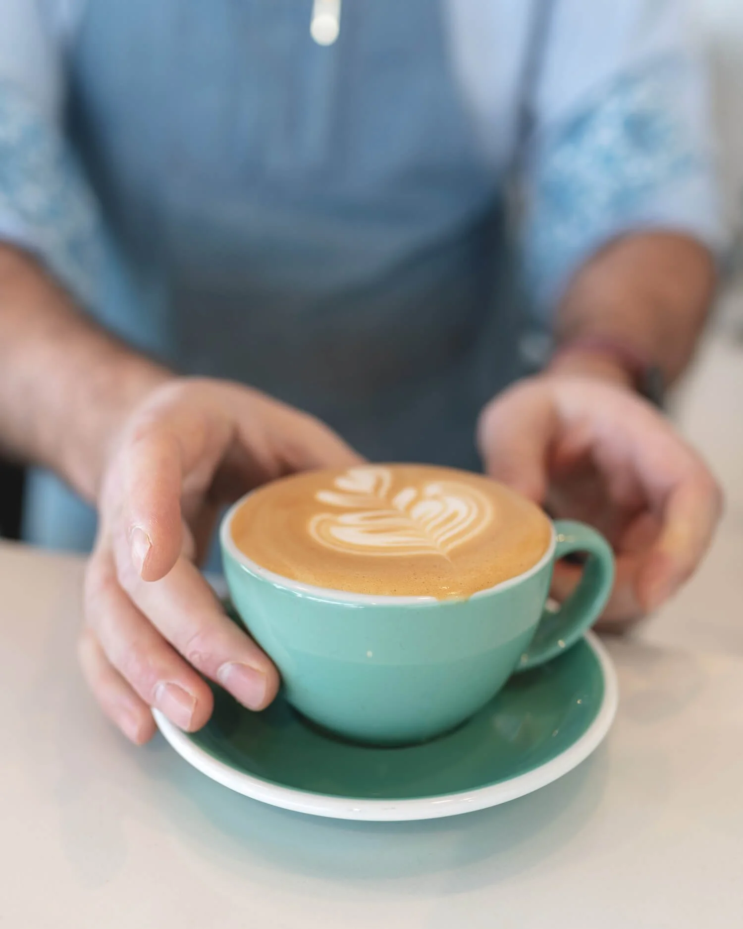 A latte with rosetta art in a teal cup and saucer, being set down on the counter