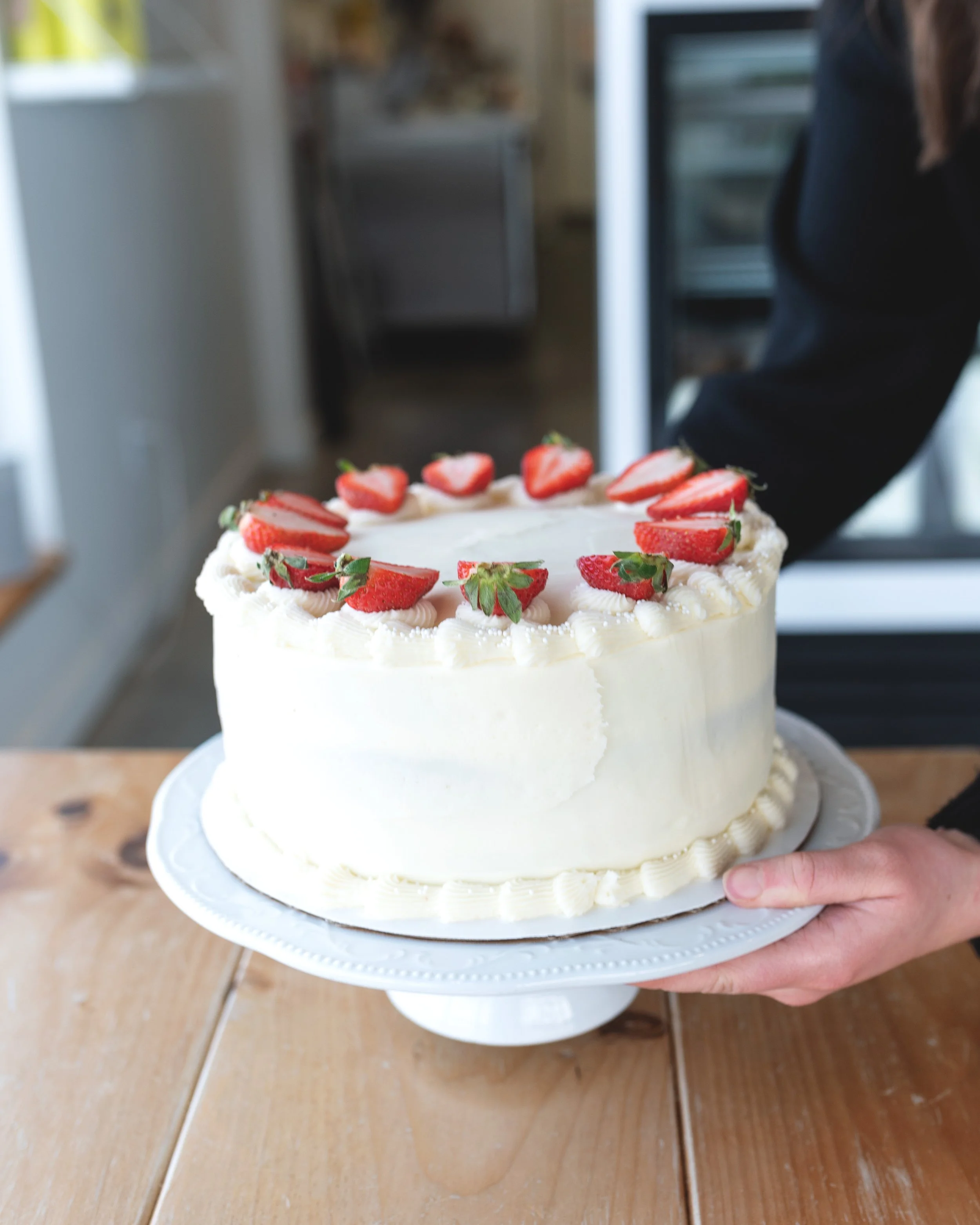 A white buttercream cake topped with fresh strawberries, held on a cake stand