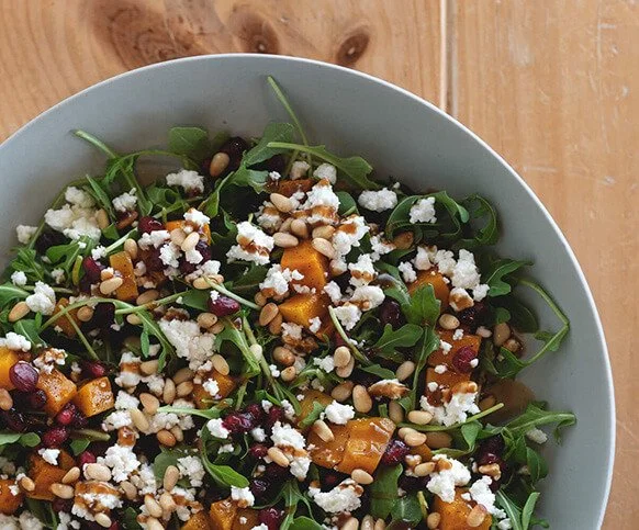 A bowl of arugula salad with roasted squash, feta, pine nuts and cranberries