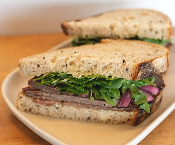 A sliced steak sandwich with greens and pickled onion on artisan bread